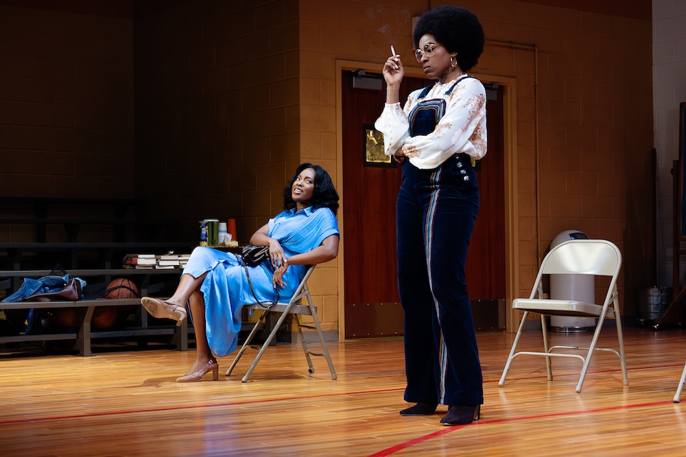 Two women in a gymnasium, one sitting in a chair, the other standing smoking a cigarette.