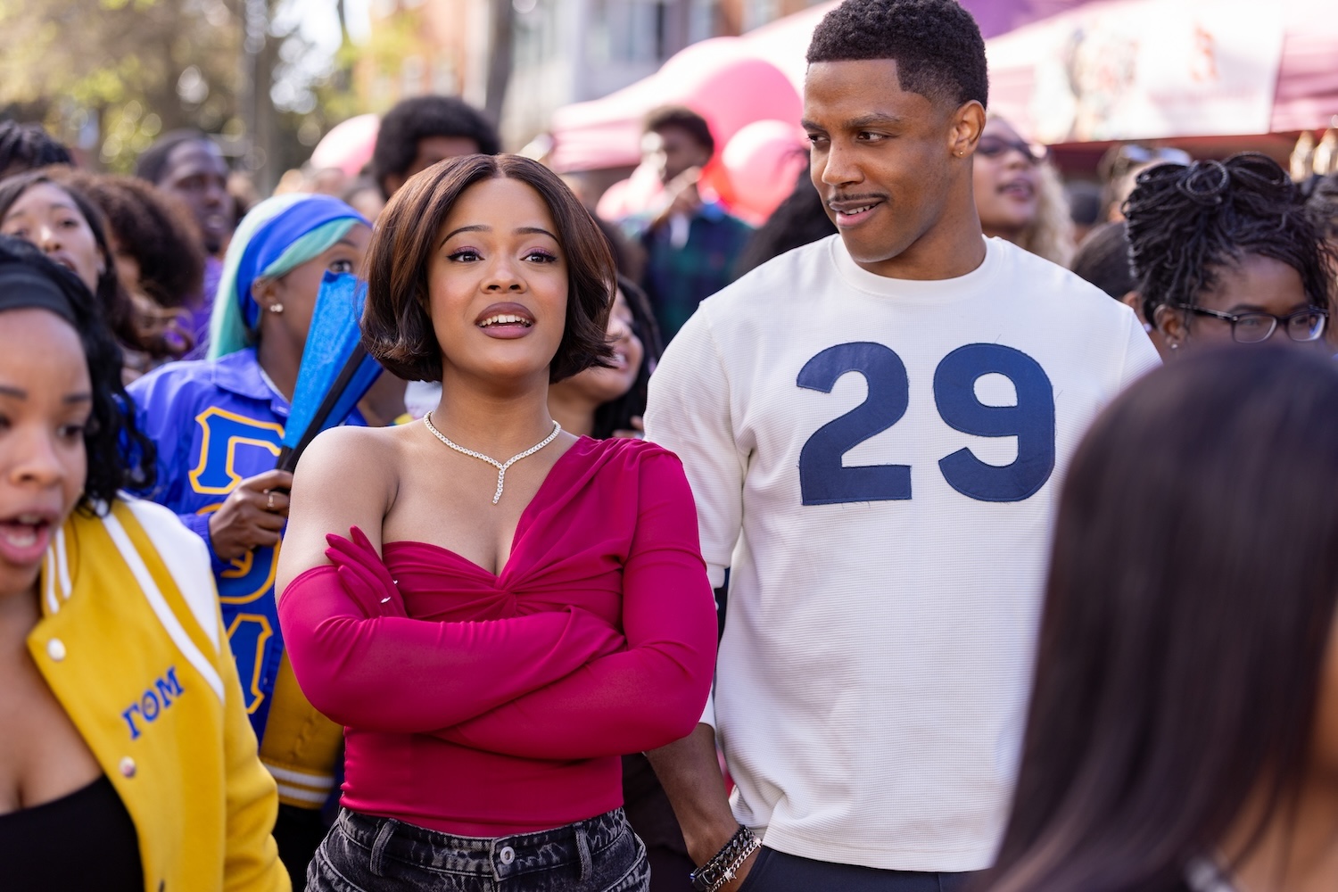 "A woman in a red top stands with her arms crossed next to a man in a white shirt with the number 29. They are outside among a diverse group of people, possibly at a festival or public event."